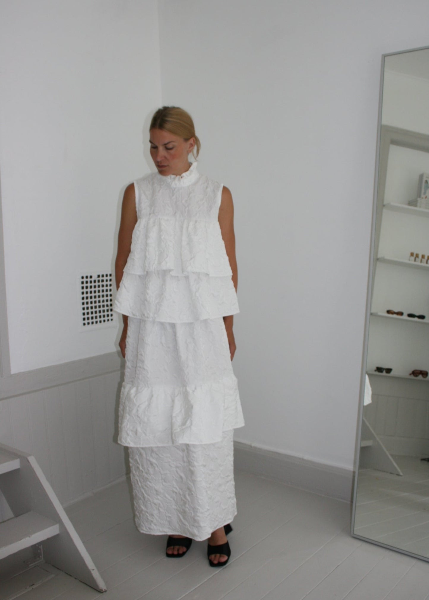 Woman wearing a white dress standing in a minimalistic room with a mirror.