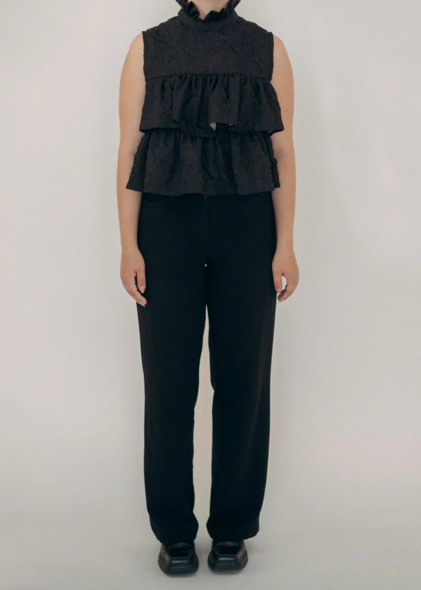 Person wearing a black sleeveless ruffled top and black pants on a white background