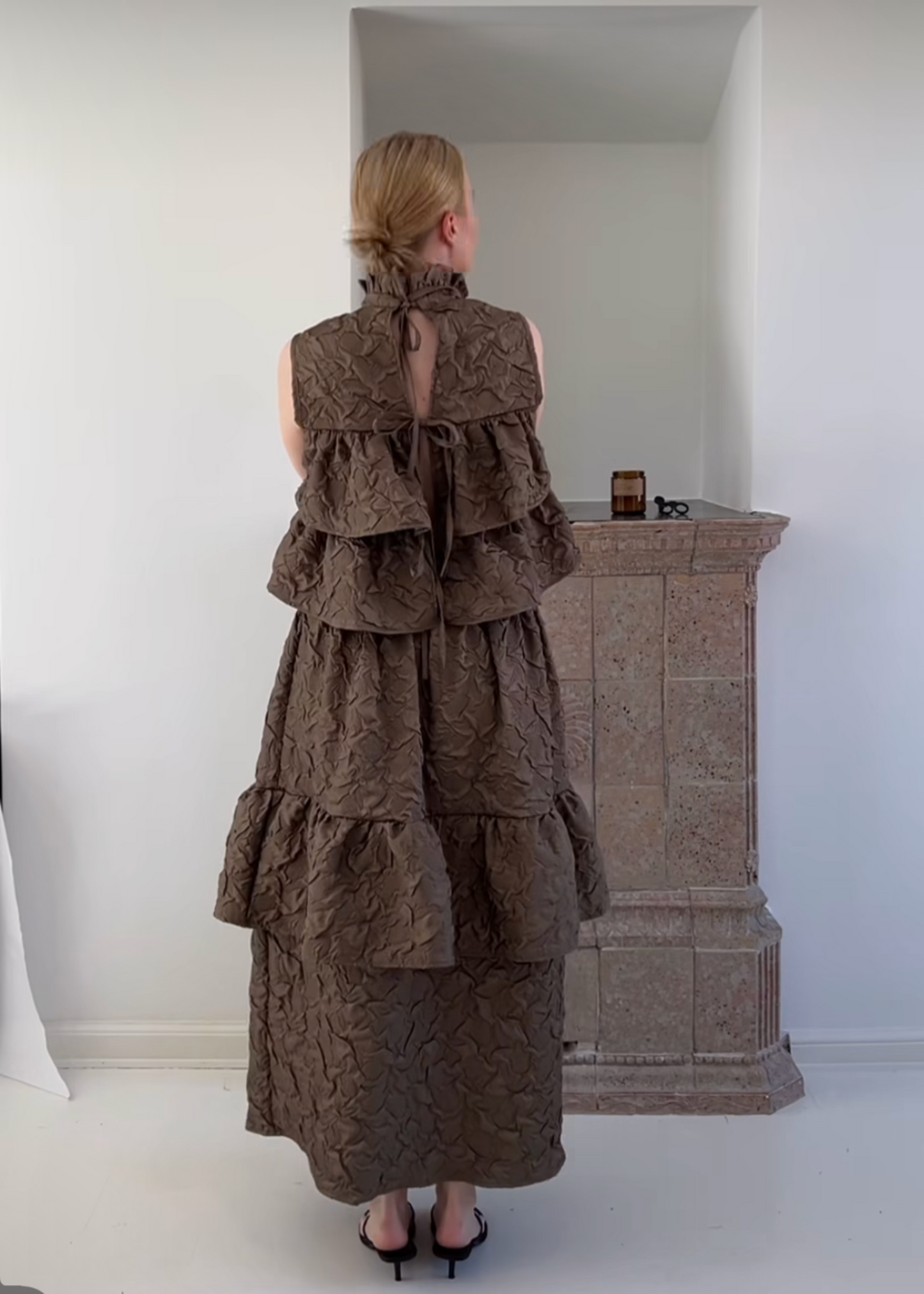 Woman wearing a long, brown, ruffled dress in a minimal white room.
