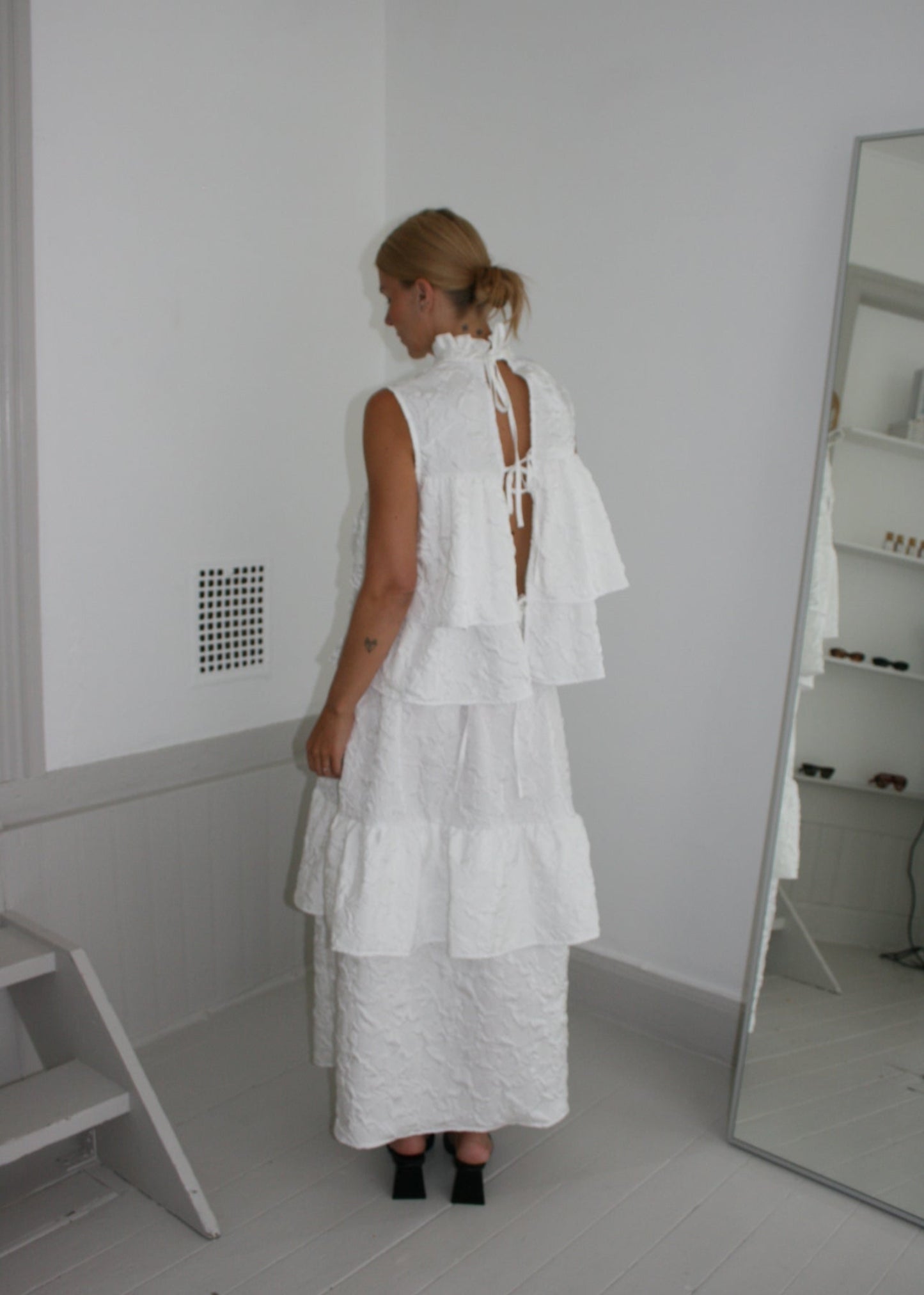 Person wearing a white dress standing on a white floor with a mirror in the background.