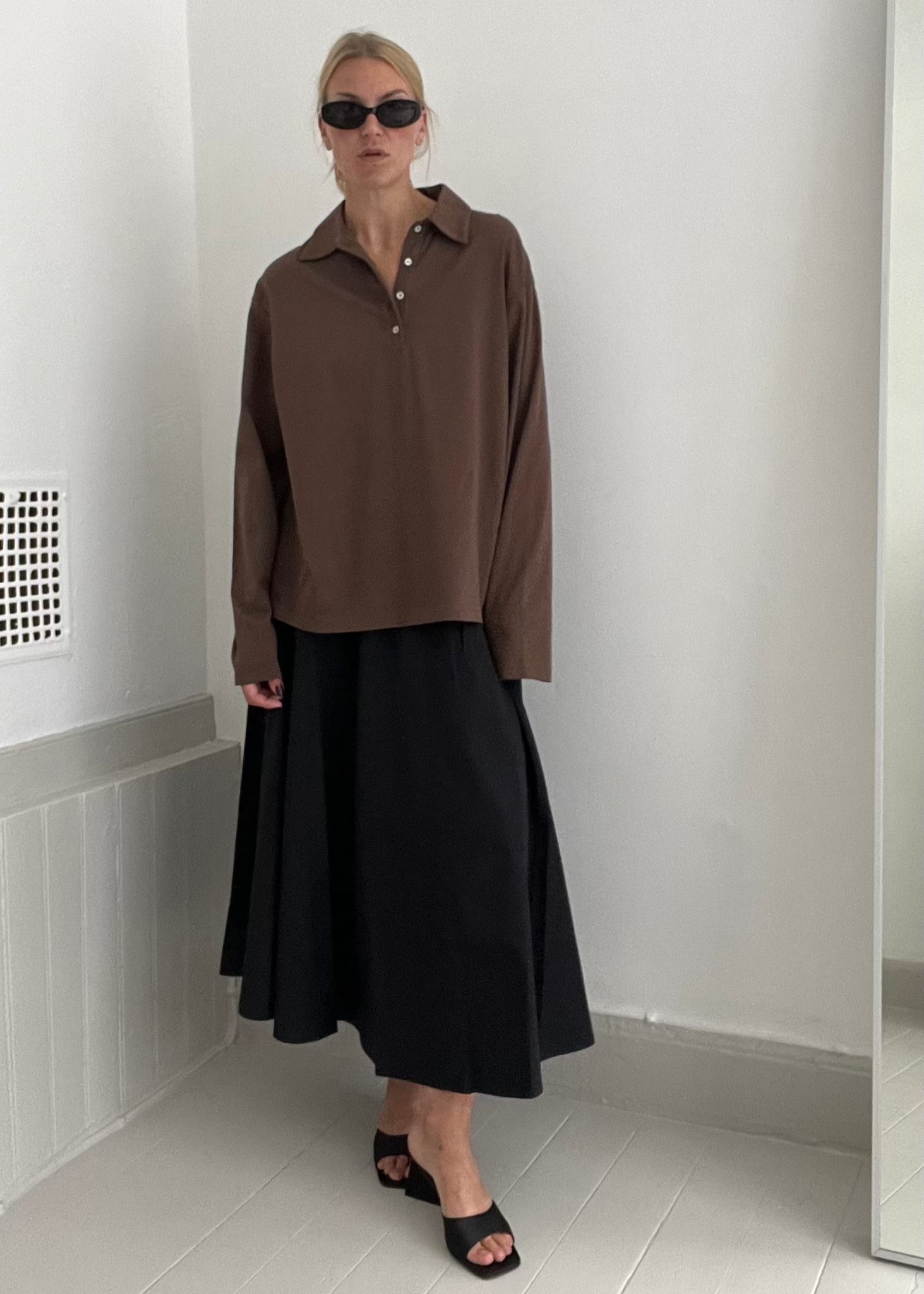 Person wearing a brown shirt and black skirt standing in a room with a white wall and floor.