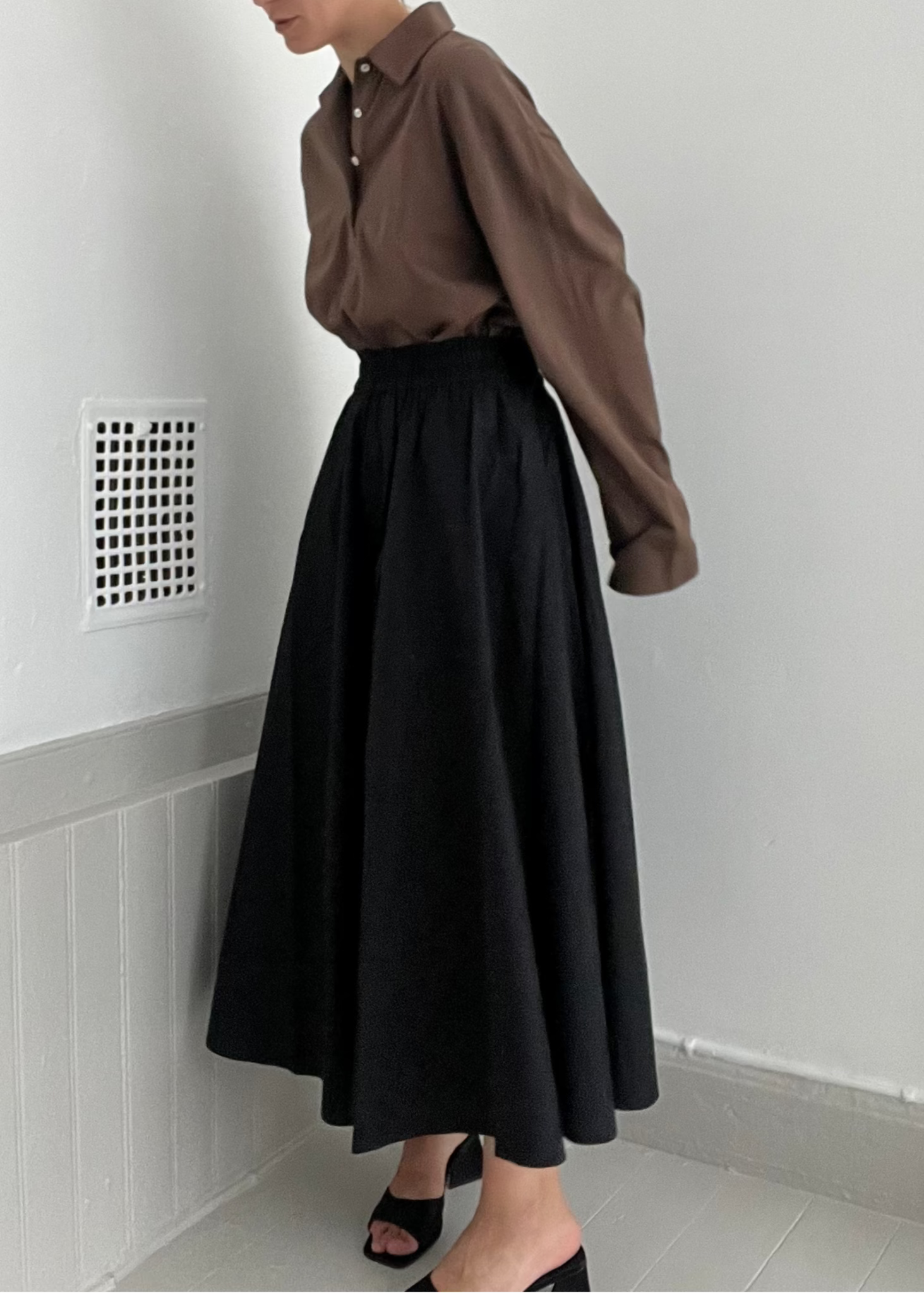 Person wearing a brown shirt and black skirt against a white wall.