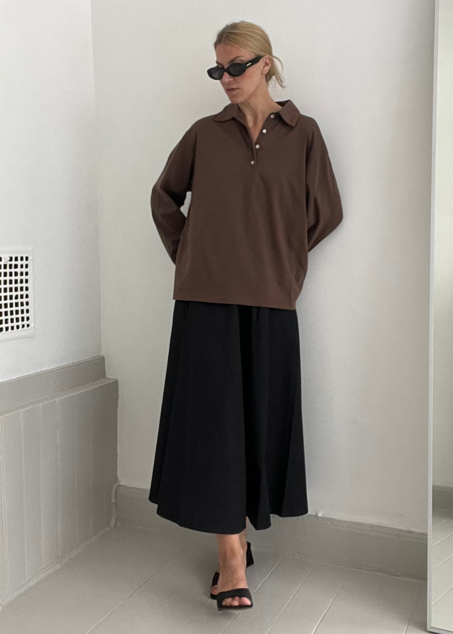 Person wearing a brown shirt and black pants standing against a white wall.