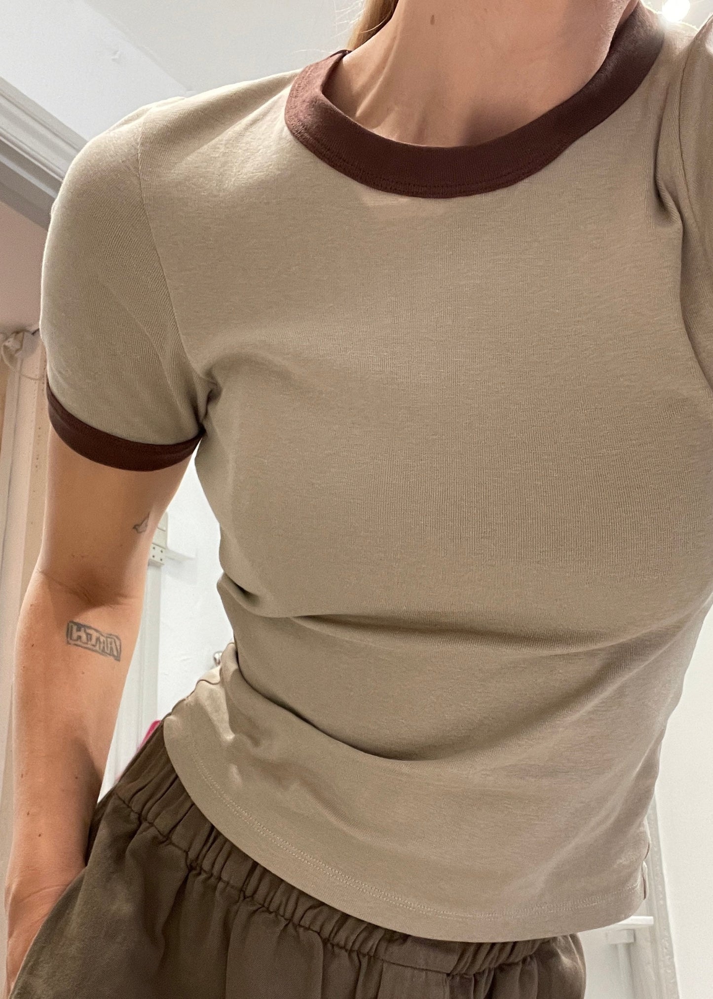 Person wearing a beige t-shirt with brown trim and brown pants indoors.