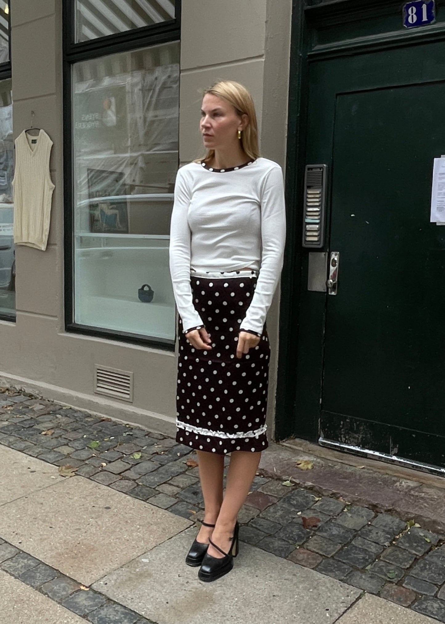 Woman in a white long-sleeve top and black polka dot skirt standing on a sidewalk.