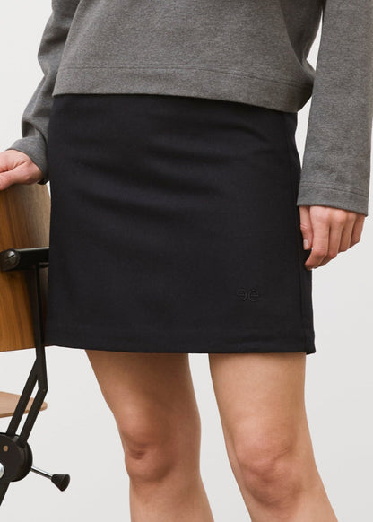 Woman wearing a gray sweater and black skirt with a striped scarf, standing next to a wooden chair against a white wall.