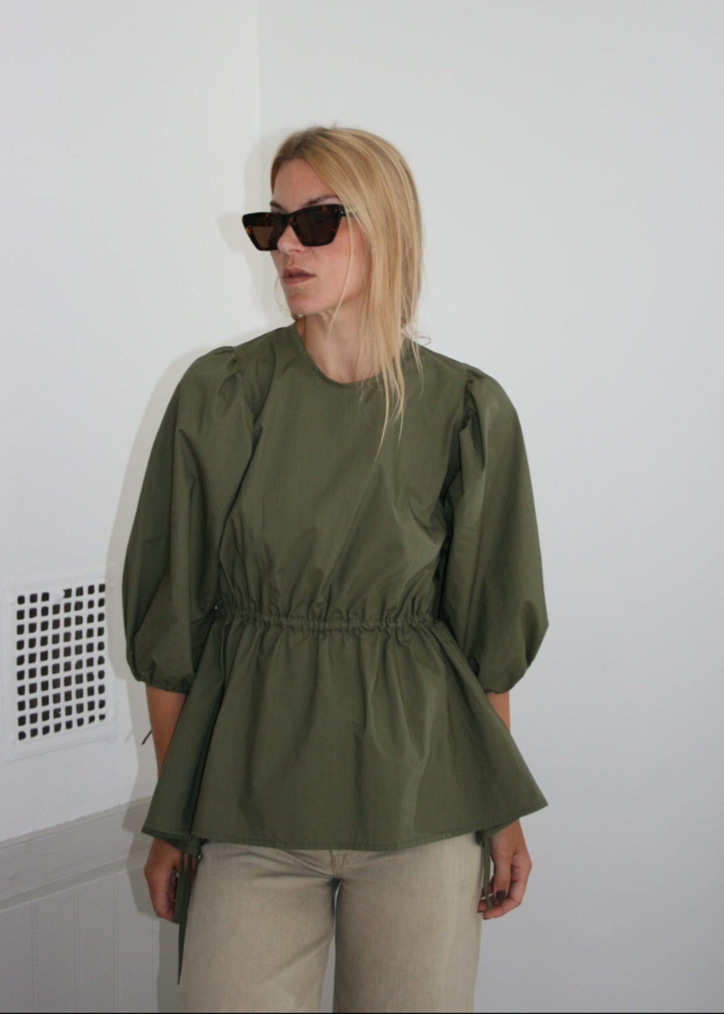 Person wearing a mette bjerg studio green blouse and sarth sunglasses on a white floor with a white wall background
