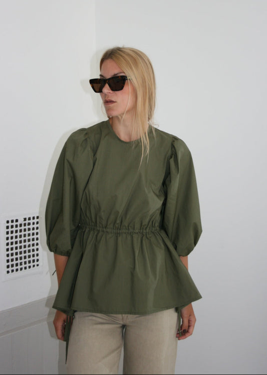 Person wearing a mette bjerg studio green blouse and sarth sunglasses on a white floor with a white wall background