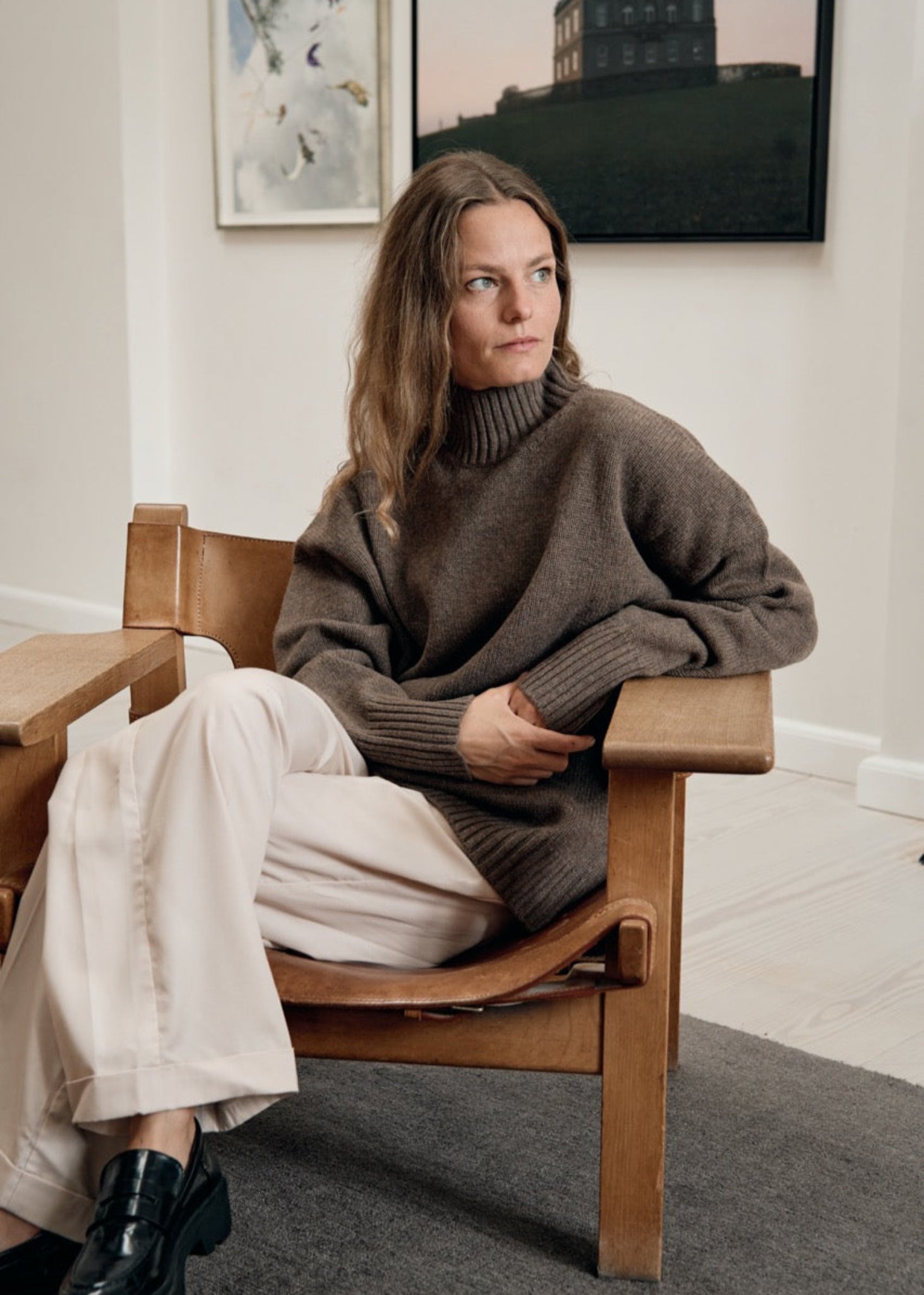 Woman sitting on a wooden chair wearing a brown sweater and white pants in a room with framed pictures on the wall.