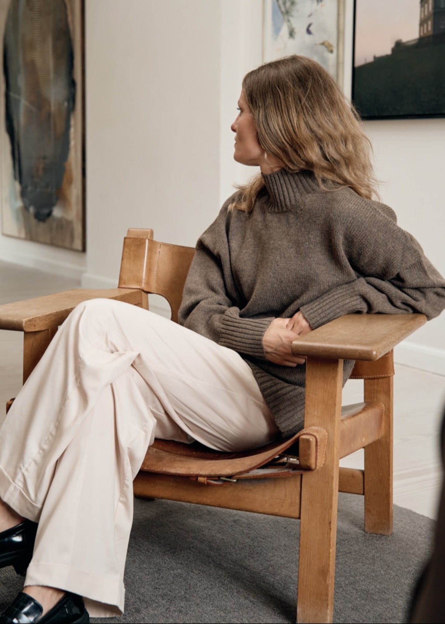 Woman sitting on a wooden chair in a room with framed pictures on the wall.