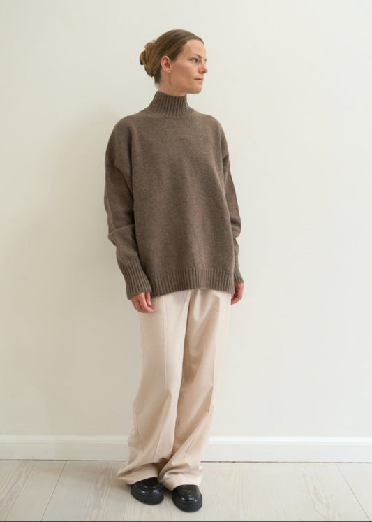 Person wearing a brown sweater and beige pants against a white wall