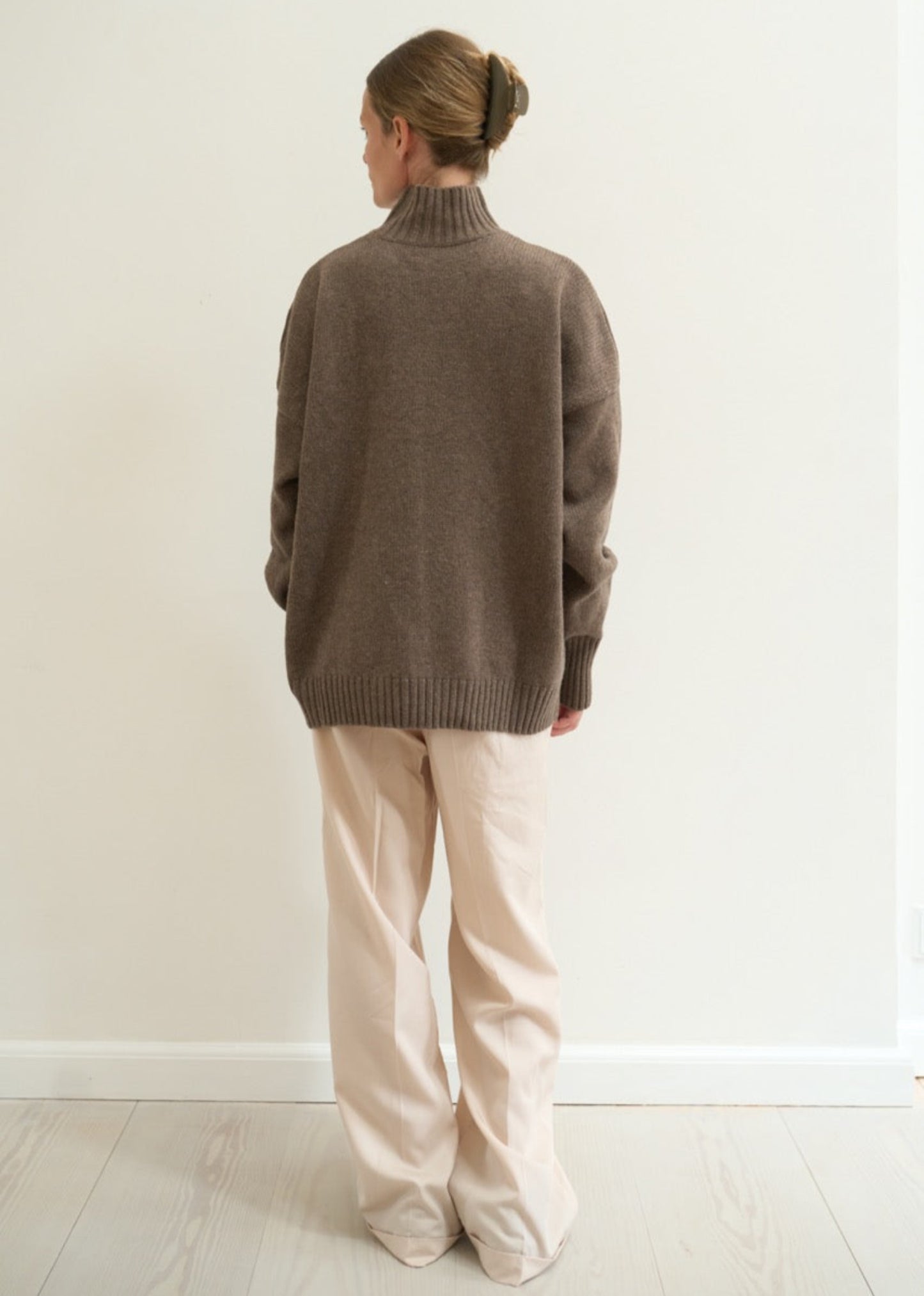 Person wearing a brown sweater and beige pants against a white wall