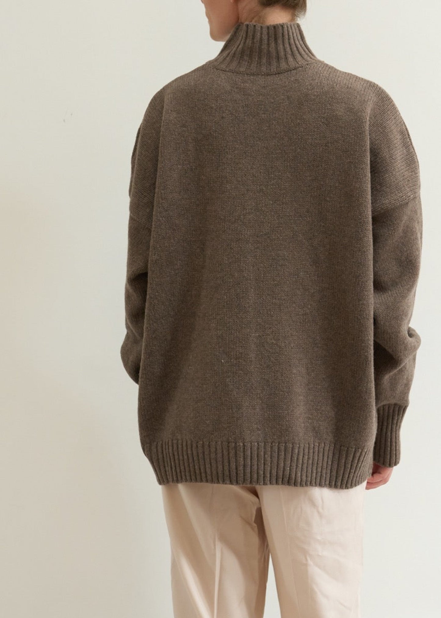 Person wearing a brown sweater and beige pants against a plain background