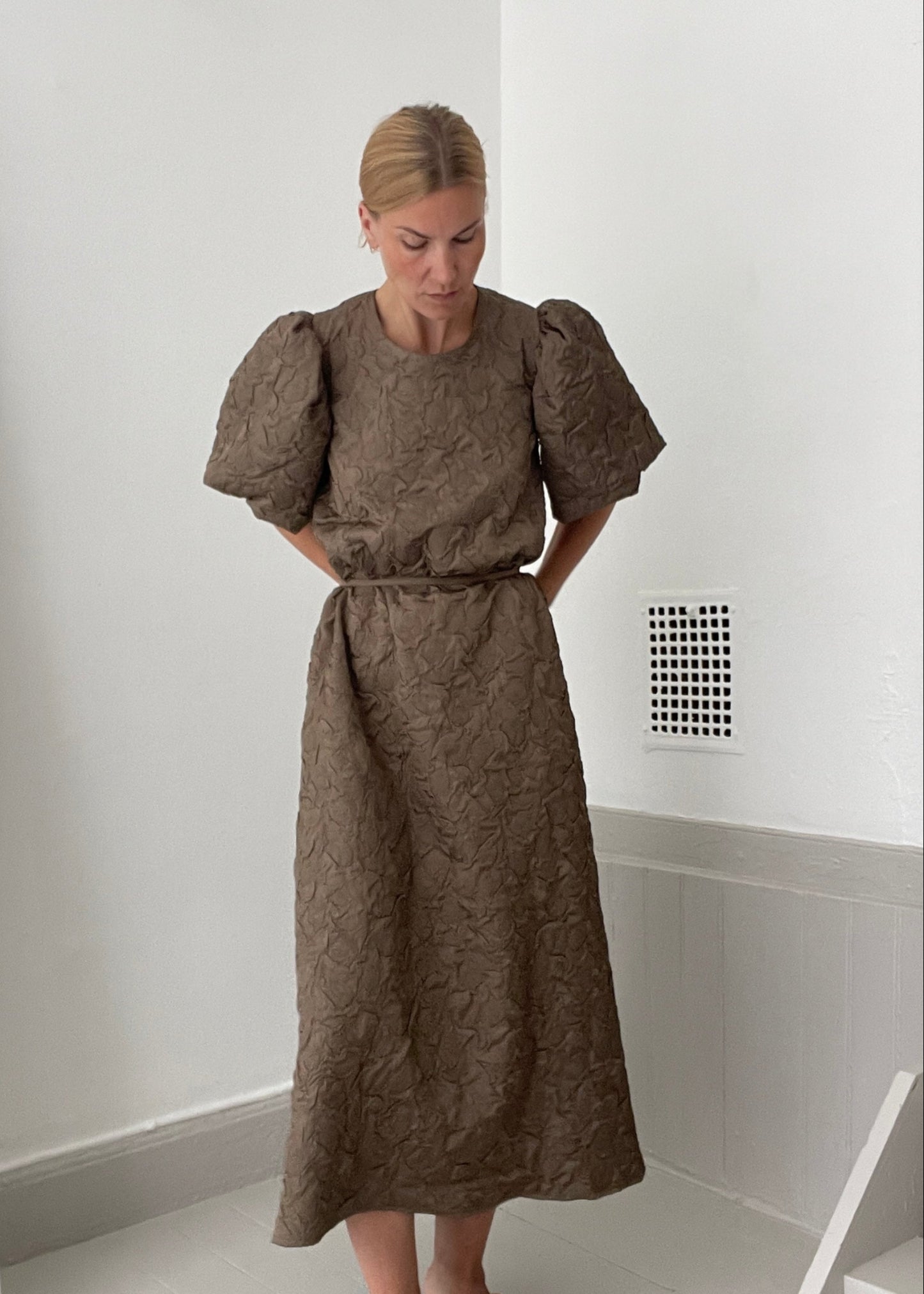 Woman wearing a brown dress with puffed sleeves in a minimal indoor setting.