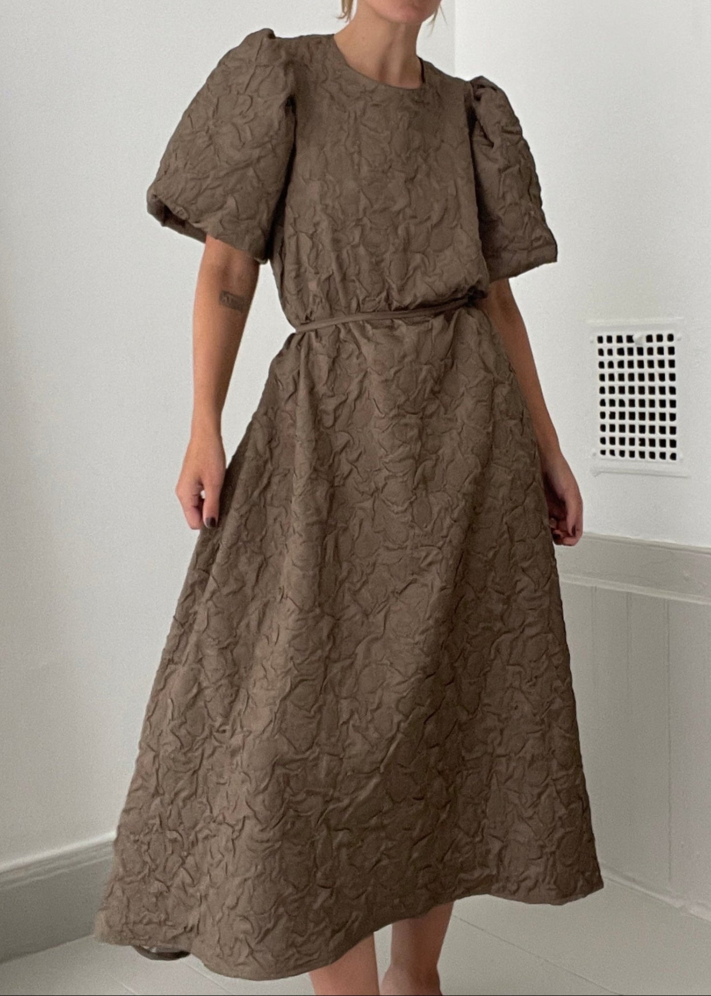 Woman wearing a brown dress with puff sleeves in a minimal indoor setting.