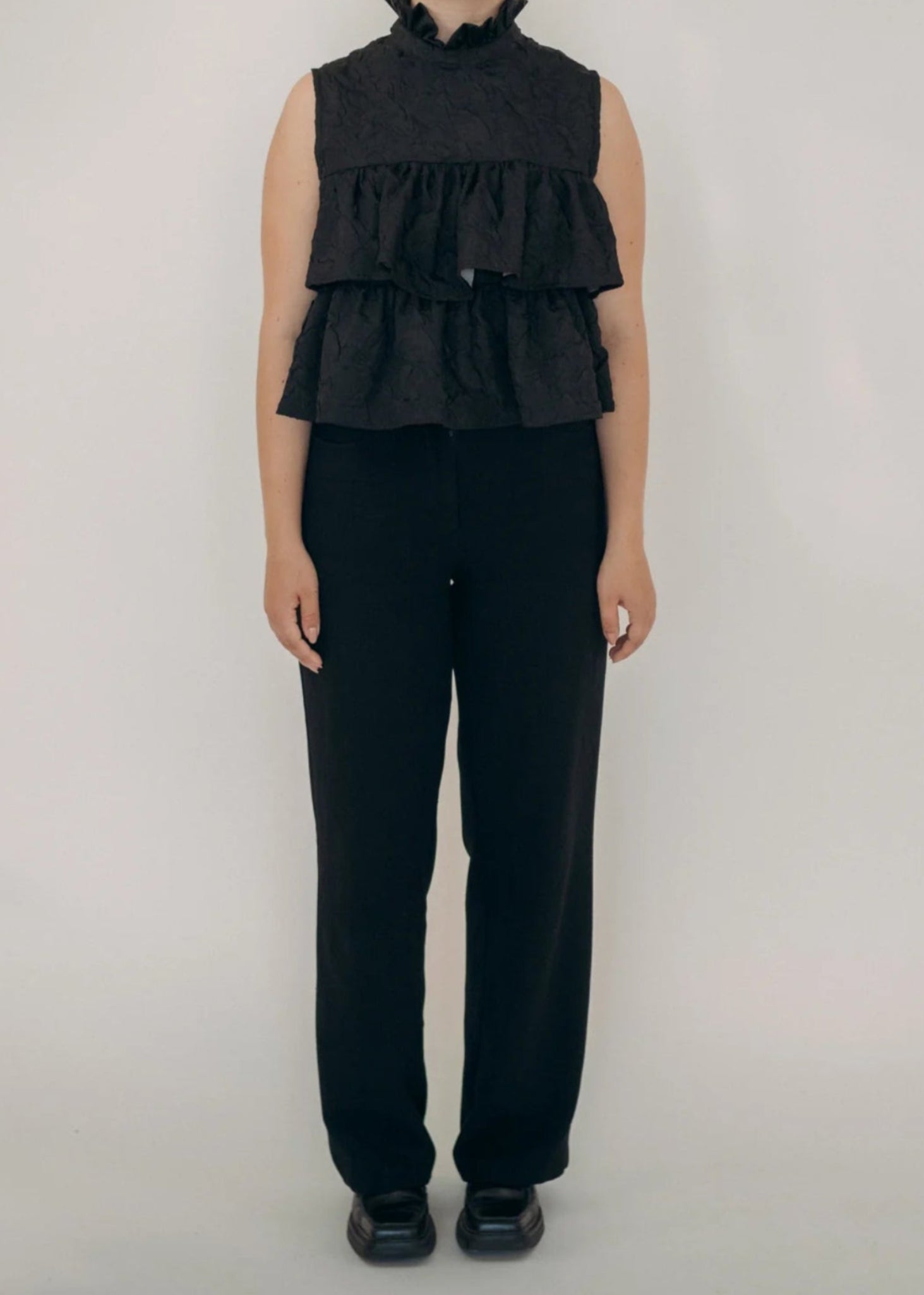 Person wearing a black sleeveless ruffled top and black pants on a white background
