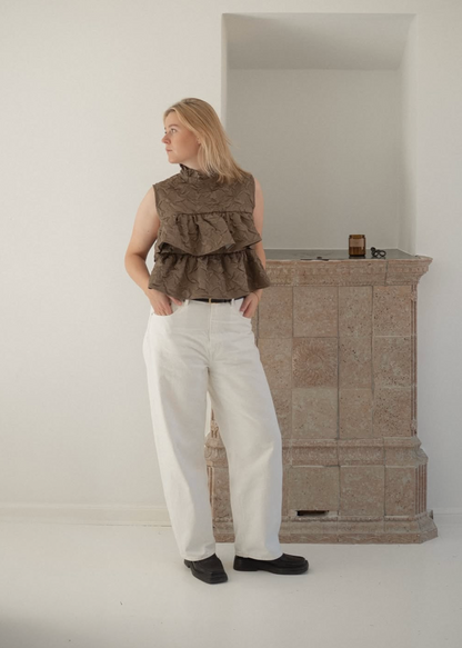 Person wearing a brown vest and white pants standing in a minimalistic room with a wooden cabinet.