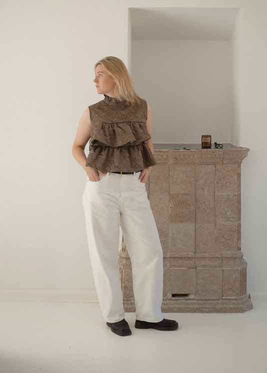 Person wearing a brown vest and white pants standing in a minimalistic room with a wooden cabinet.