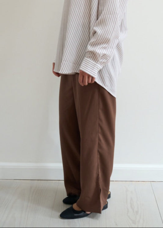 Person wearing a white shirt and brown pants standing against a plain white wall.