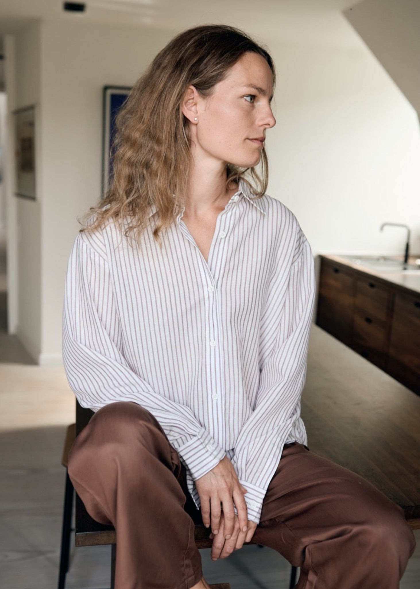 Woman sitting on a chair wearing a striped shirt and brown pants in an indoor setting.