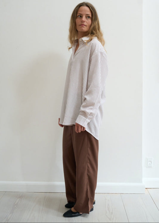Woman wearing a white striped shirt and brown pants against a white wall.