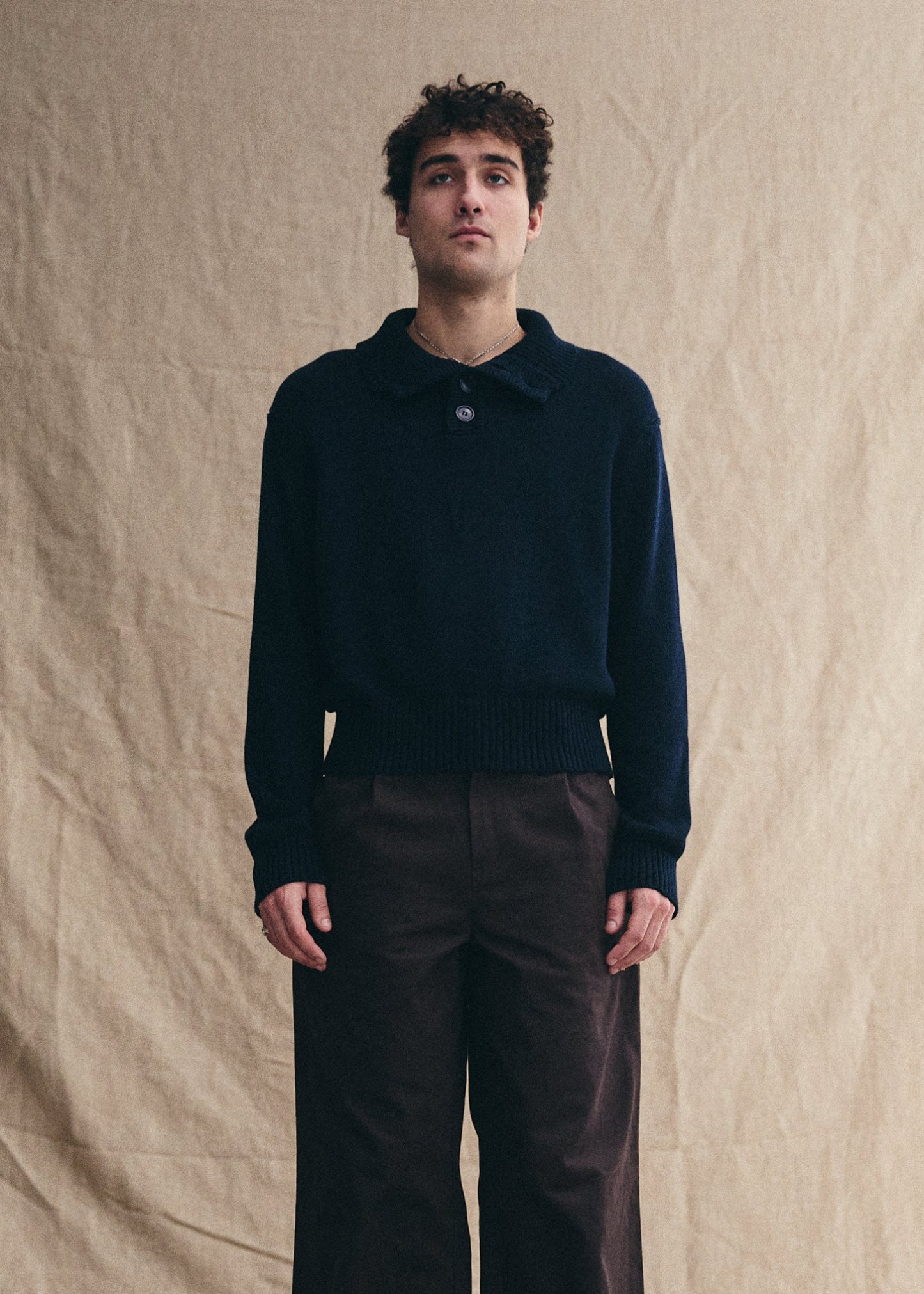 Man wearing a dark sweater and brown pants against a beige backdrop