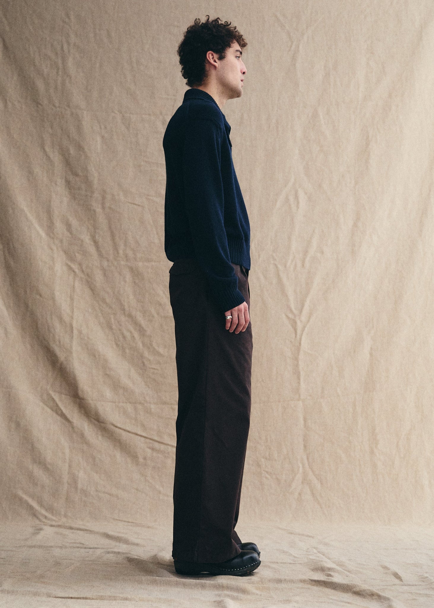 Man standing against a beige backdrop wearing a dark sweater and pants.