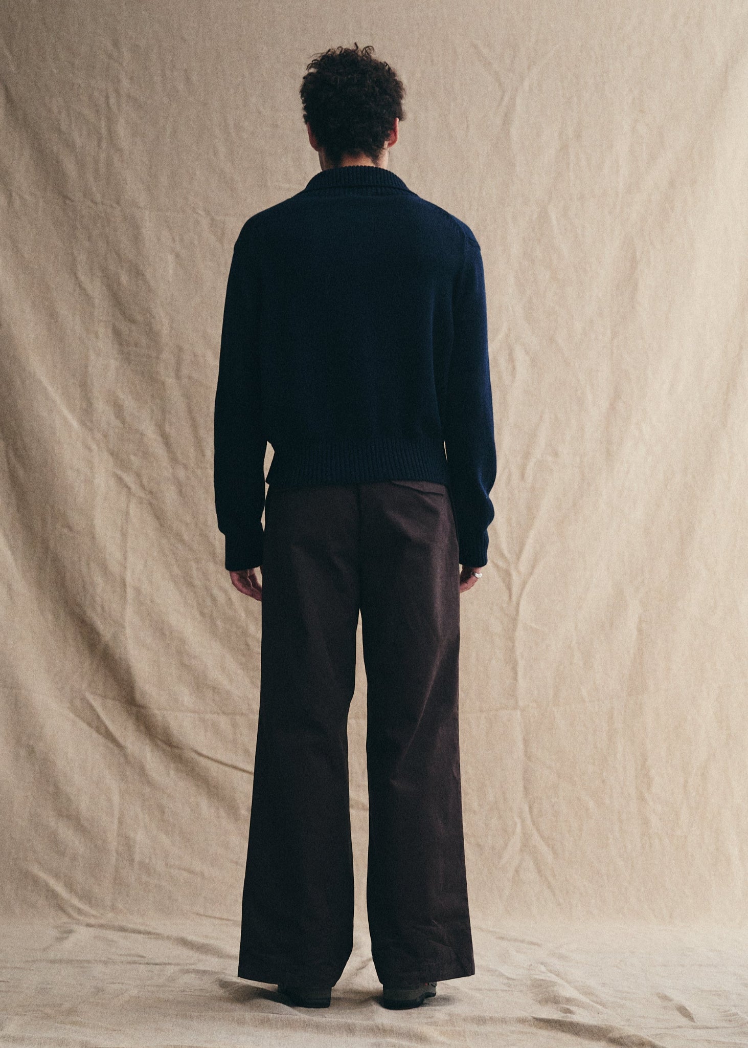 Person wearing a dark sweater and brown pants standing against a beige backdrop