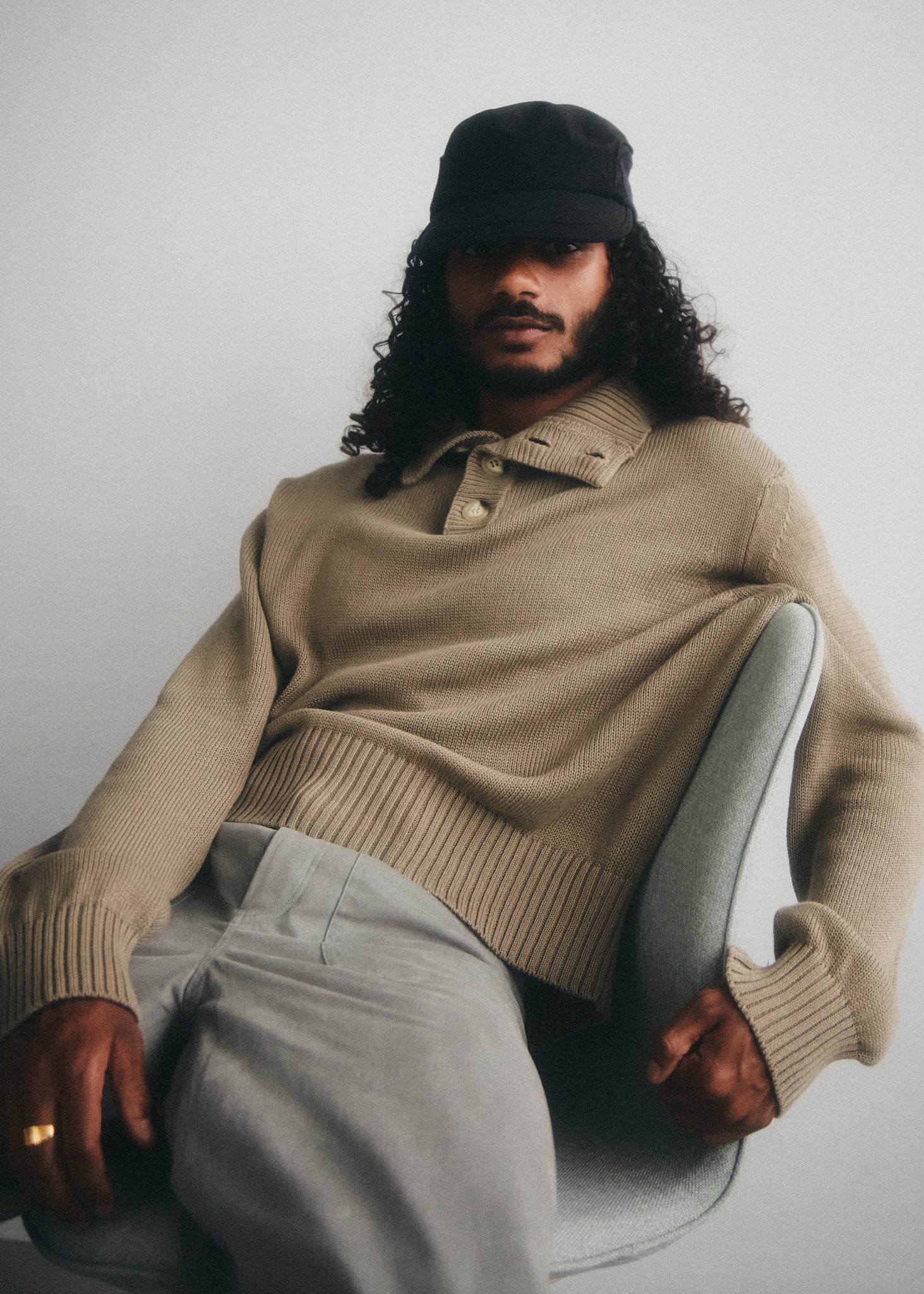 Man wearing a beige sweater and black cap sitting on a white surface.