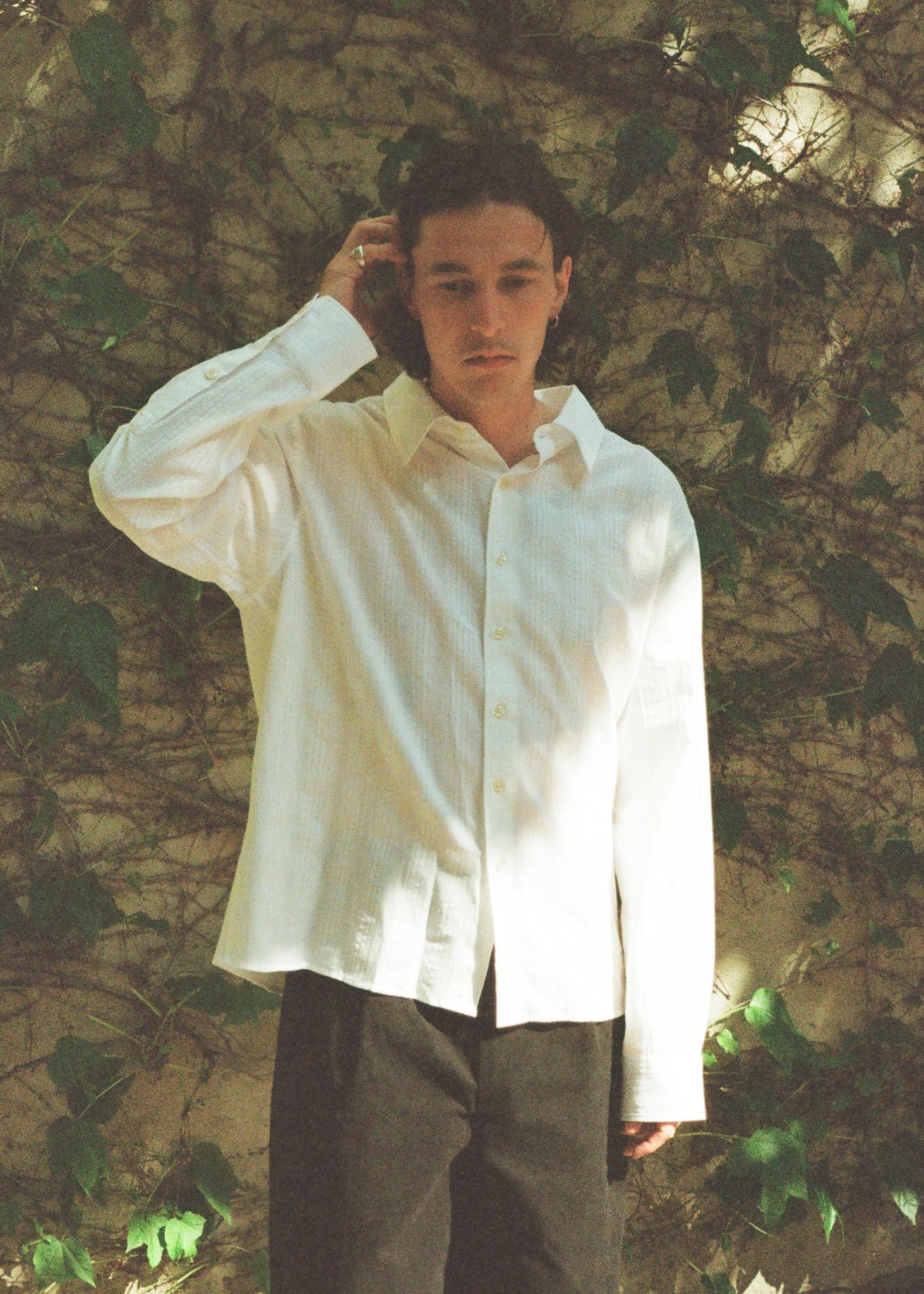 Person wearing a white shirt and dark pants standing against a textured wall with greenery.
