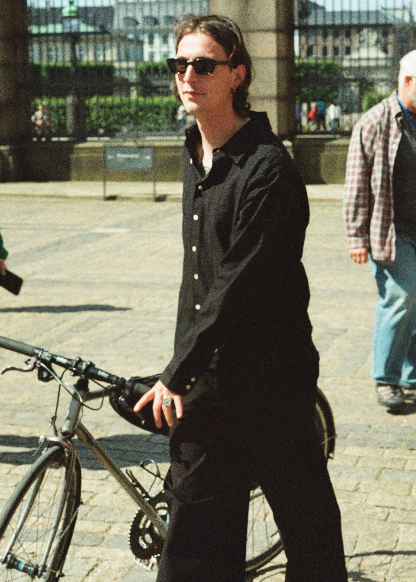 Person in black outfit with sunglasses standing next to a bicycle on a city street.