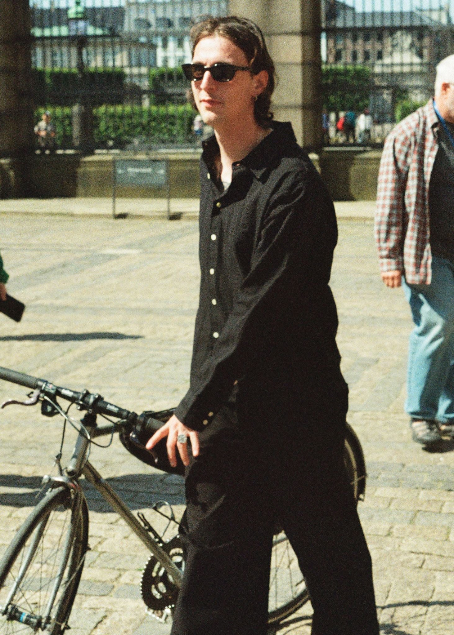 Person in black outfit with sunglasses standing next to a bicycle on a city street.