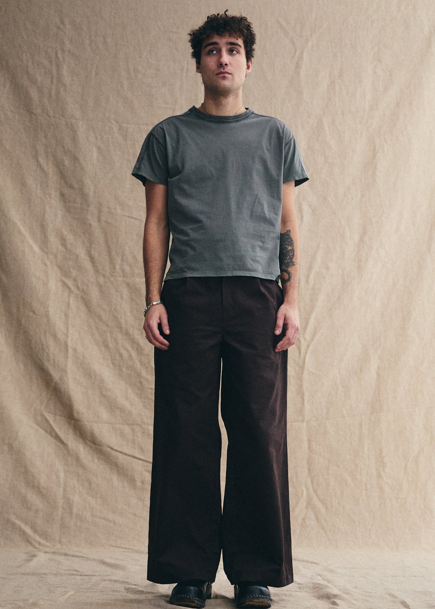 Man wearing a gray t-shirt and dark pants standing against a beige backdrop