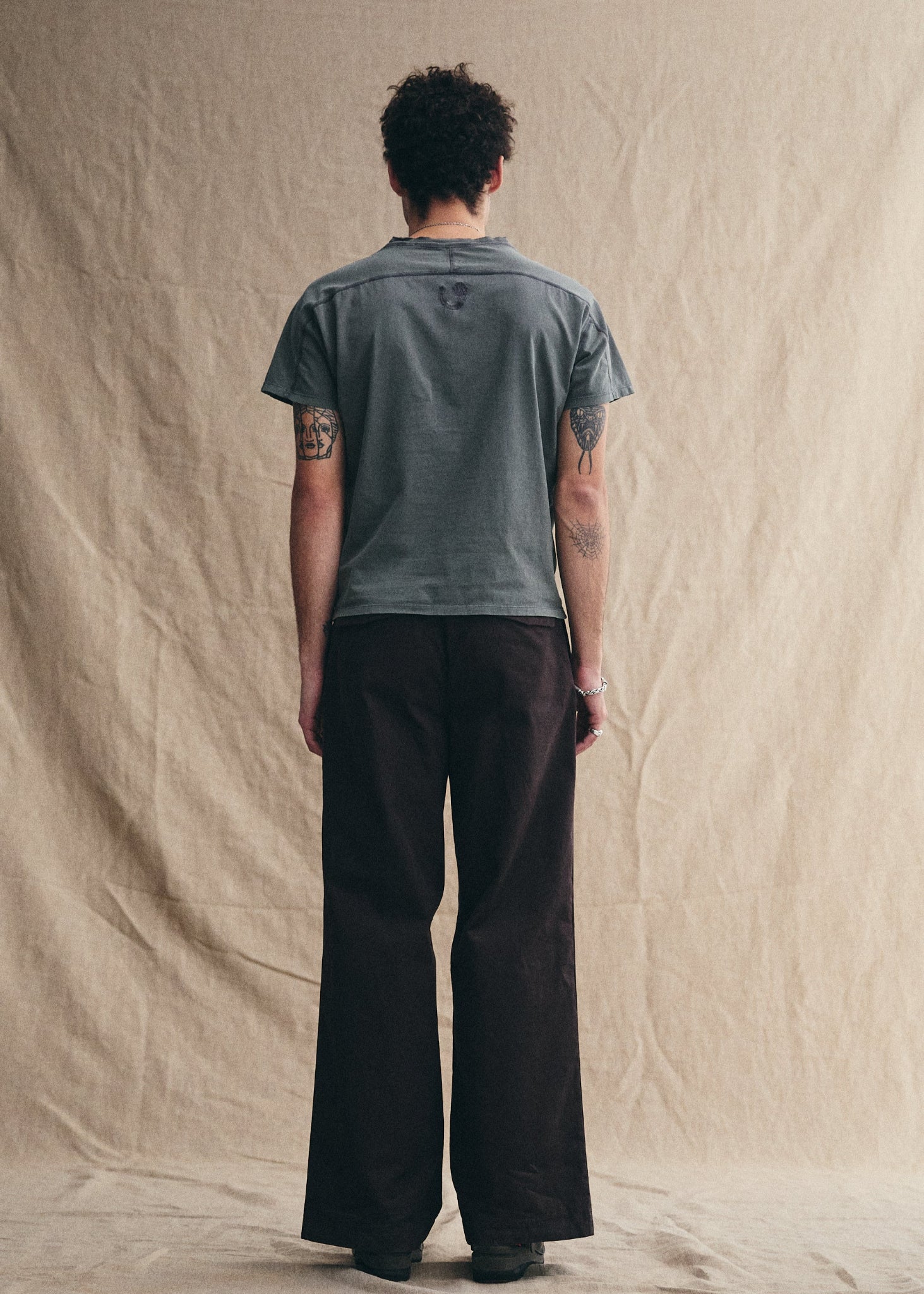 Person wearing a gray t-shirt and dark pants standing against a beige backdrop