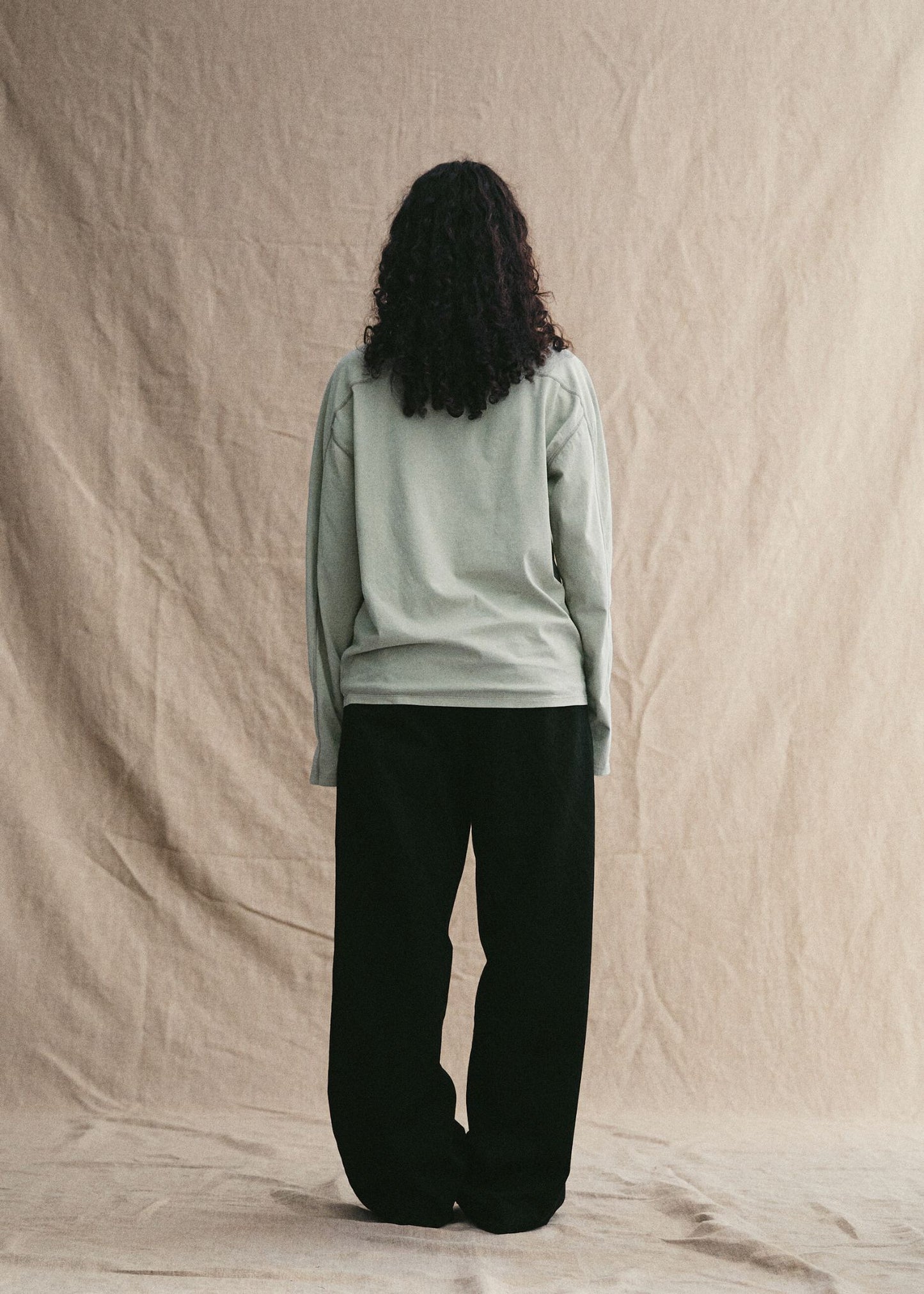 Person wearing a light green long-sleeve shirt and black pants against a beige background