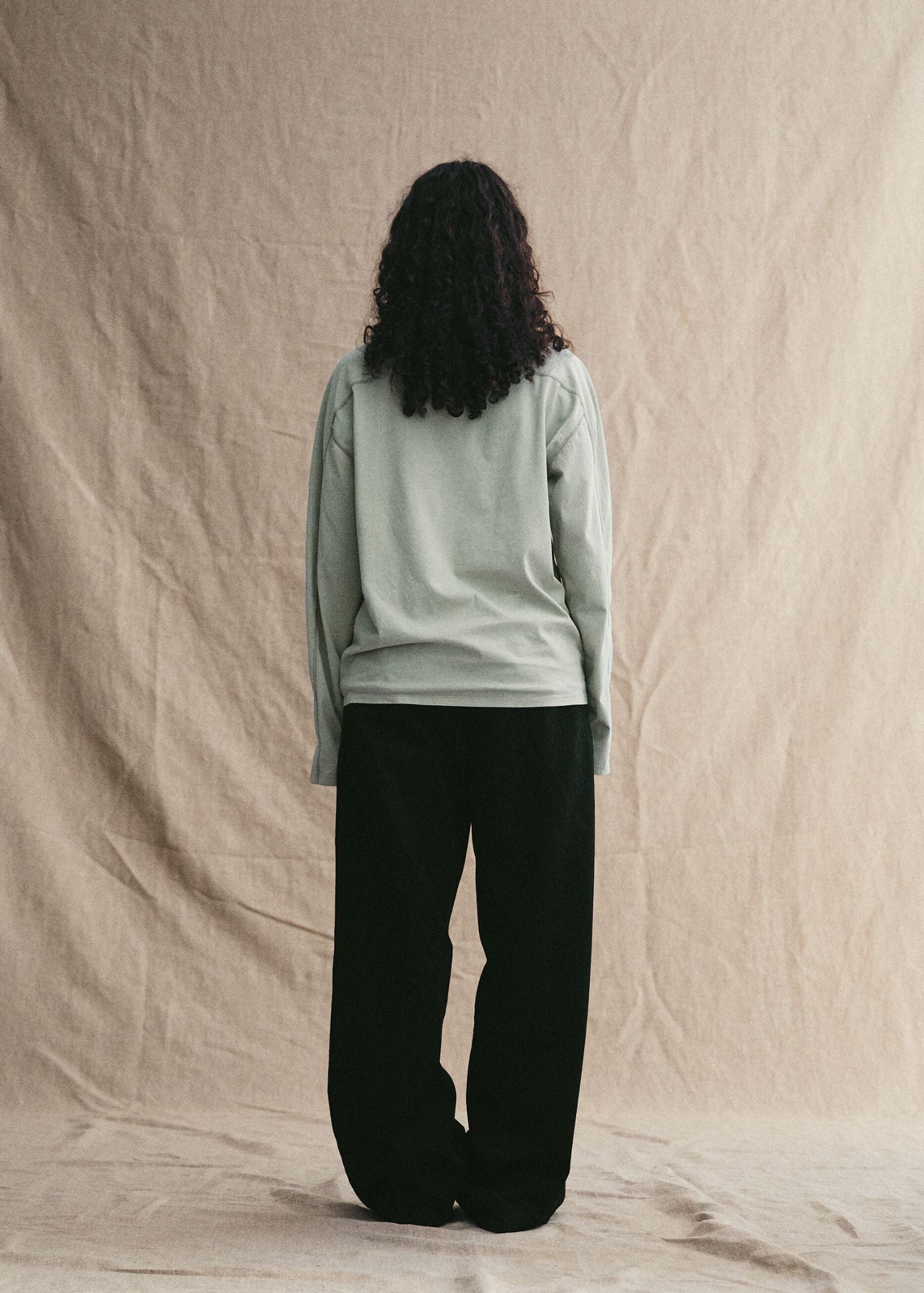 Person wearing a light green long-sleeve shirt and black pants against a beige background
