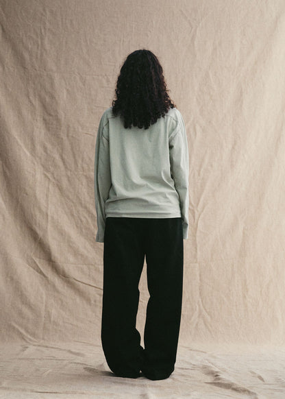 Person wearing a light green long-sleeve shirt and black pants against a beige background